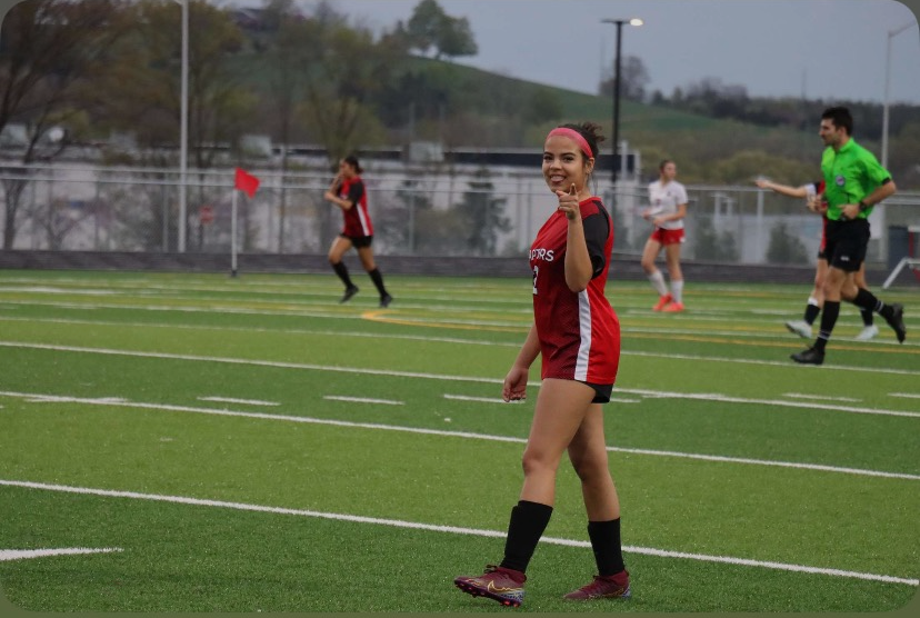 Freshman Emily Barahona stands on the field smiling and pointing towards the camera.